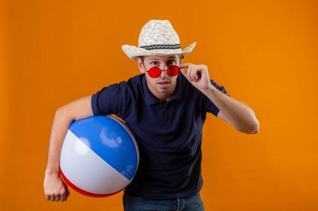 Junger hübscher Mann im Sommerhut, der aufblasbaren Ball hält, der Kamera betrachtet, die Brille abnimmt, als Blick auf Kamera überrascht und fasziniert steht über orange Hintergrund