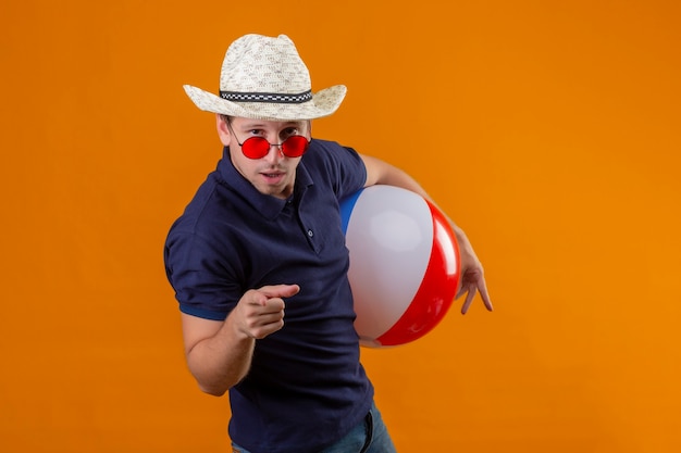 Kostenloses Foto junger hübscher mann im sommerhut, der rote sonnenbrille hält, die aufblasbaren ball zeigt, der mit dem finger zur kamera mit dem sicheren blick zeigt, der über orange hintergrund steht