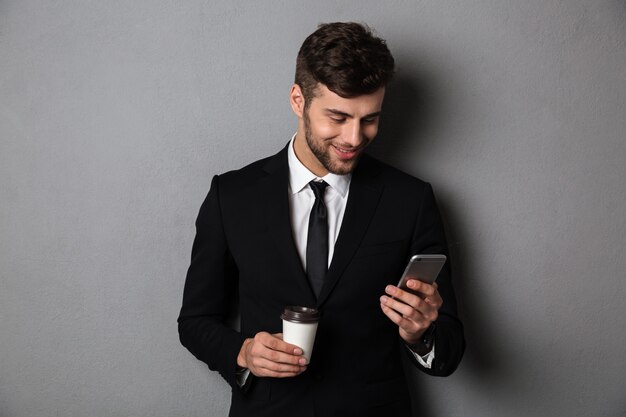 Junger hübscher Mann in der formellen Abnutzung, die Nachrichten auf Smartphone überprüft, während Kaffee zum Mitnehmen hält