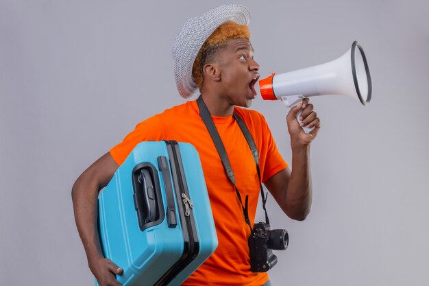 Junger hübscher reisender Junge im Sommerhut, der orange T-Shirt hält, das Reisekoffer hält, der zum Megaphon schreit, das über weißer Wand steht