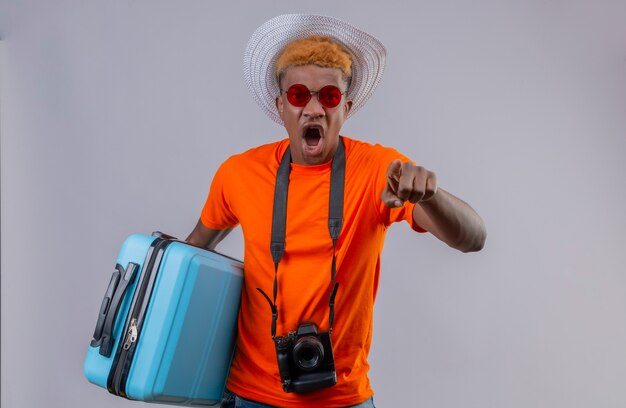 Junger hübscher reisender Junge im Sommerhut, der orange T-Shirt hält, das Reisekoffer hält, der zur Kamera mit dem Finger zeigt, der mit wütendem Gesicht schreit