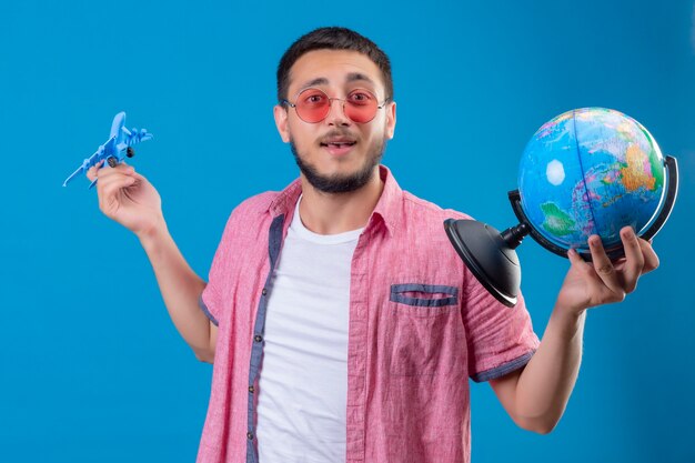 Junger hübscher reisender Kerl, der das Tragen der Sonnenbrille hält, das Spielzeugflugzeug und den Globus hält, der positiv und glücklich lächelnd freundlich steht, das über blauem Hintergrund steht