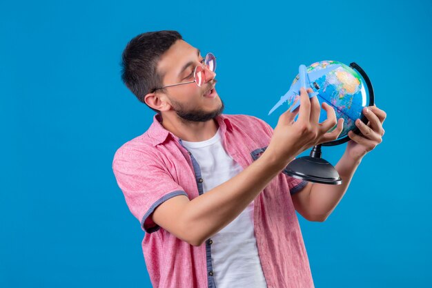 Junger hübscher reisender Kerl, der das Tragen der Sonnenbrille hält, die Spielzeugflugzeug und Globus hält, die glückliches und positives Stehen über blauem Hintergrund spielen