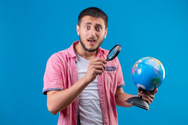 Junger hübscher reisender Kerl, der Globus hält und ihn unter Verwendung der Lupe betrachtet, die überrascht steht, über blauem Hintergrund stehend