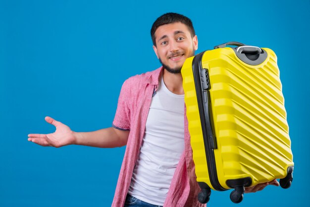 Junger hübscher reisender Kerl, der Koffer hält, der verwirrendes lächelndes Stehen mit erhöhter Hand über blauem Hintergrund schaut