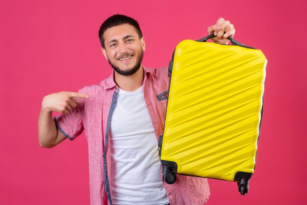 Junger hübscher reisender Kerl, der Koffer hält Kamera mit Lächeln auf Gesicht zeigend mit Finger auf Koffer steht über rosa Hintergrund