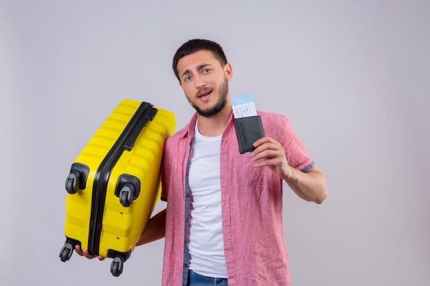 Junger hübscher reisender Kerl, der Koffer und Flugtickets hält Kamera mit selbstbewusstem Lächeln auf Gesicht glücklich und positiv stehend über weißem Hintergrund betrachtet