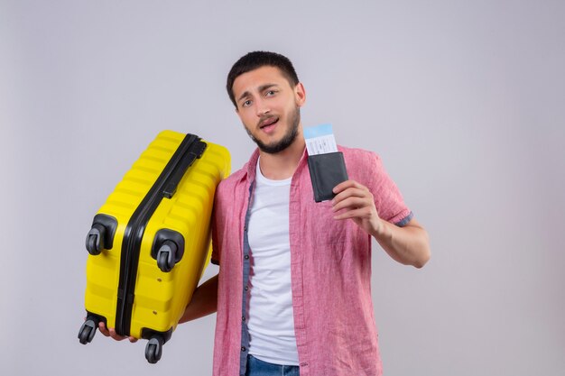 Junger hübscher reisender Kerl, der Koffer und Flugtickets hält Kamera mit selbstbewusstem Lächeln auf Gesicht glücklich und positiv stehend über weißem Hintergrund betrachtet