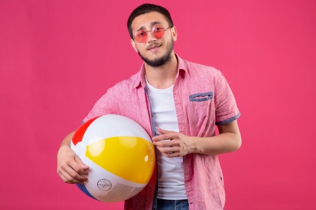 Junger hübscher reisender Kerl, der Sonnenbrille hält, die aufblasbaren Ball betrachtet Kamera mit sicherem Lächeln auf Gesicht steht, das über rosa Hintergrund steht