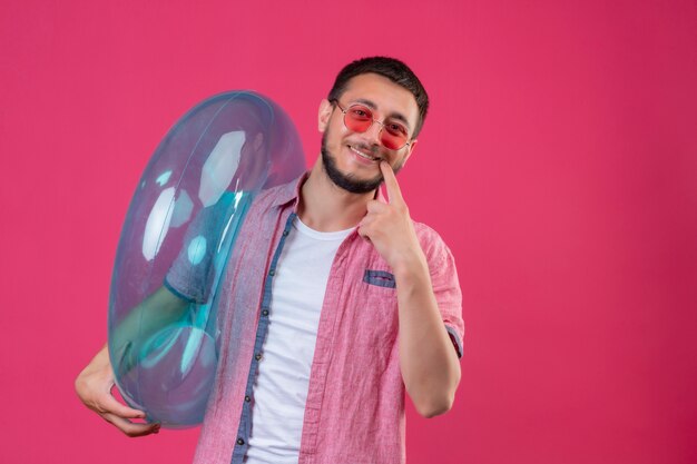 Junger hübscher reisender Kerl, der Sonnenbrille hält, die aufblasbaren Ring hält, der Kamera schaut, die fröhlich mit dem Finger auf seinen Mund zeigt, der über rosa Hintergrund steht