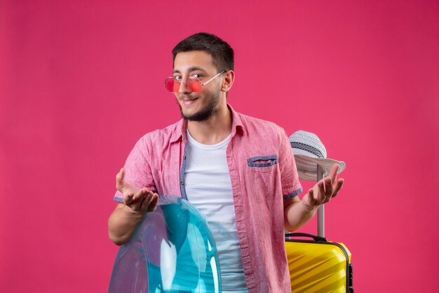 Junger hübscher reisender Kerl, der Sonnenbrille hält, die aufblasbaren Ring hält, der mit Reisekoffer steht und Kamera betrachtet, die schlau mit Armen lächelt, die über rosa Hintergrund erhoben werden