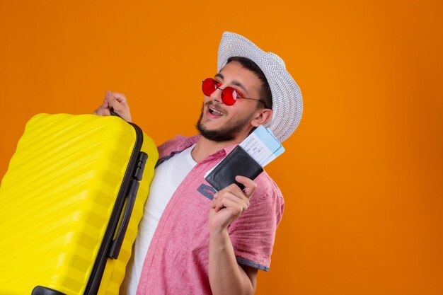 Junger hübscher Reisender Kerl, der Sonnenbrille im Sommerhut hält, der Koffer und Flugtickets hält, die zuversichtlich und glücklich lächelnd fröhlich bereit sind, über orange Hintergrund zu stehen