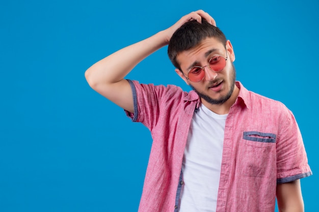 Junger hübscher reisender Kerl, der Sonnenbrille trägt, die mit Hand auf Kopf für Fehler steht, erinnert sich an Fehler schlechtes Gedächtniskonzept, das über blauem Hintergrund steht