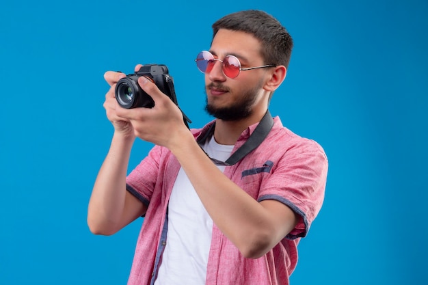 Junger hübscher reisender Kerl, der Sonnenbrillen trägt, die ein Bild mit Kamera über blauem Hintergrund stehen