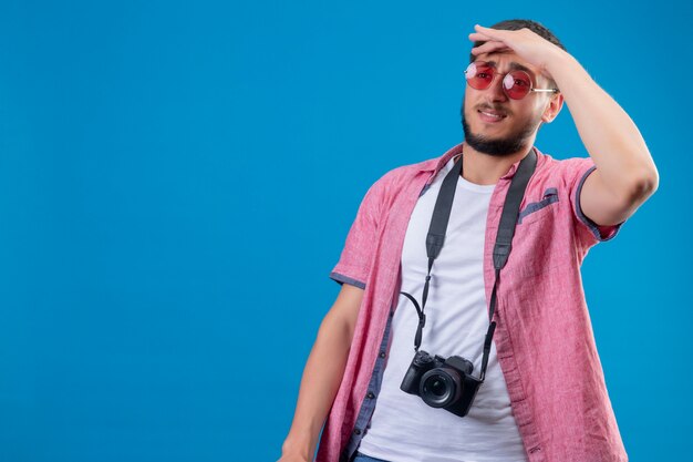 Junger hübscher Reisender Kerl mit Kamera, die Sonnenbrille trägt, die weit weg mit Hand schaut, um etwas mit verwirrendem Ausdruck zu schauen, der über blauem Hintergrund steht
