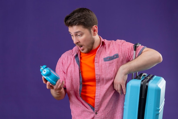 Junger hübscher reisender Mann, der blauen Koffer hält und Wecker in seiner Hand betrachtet
