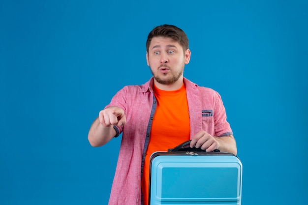 Junger hübscher reisender Mann, der Koffer hält, der mit Finger auf etwas mit Angstausdruck auf Gesicht zeigt, das über blauer Wand steht