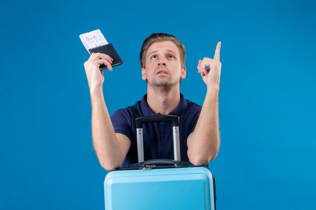 Junger hübscher reisender Mann, der Koffer und Tickets hält, die oben mit dem Finger über blauem Hintergrund stehen