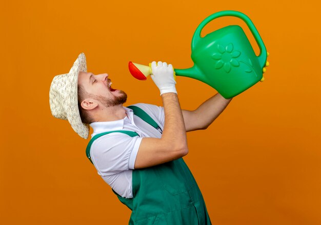 Junger hübscher slawischer Gärtner in der Uniform, die Hut und Gartenhandschuhe trinkt, die Wasser von der Bewässerungsdose isolieren