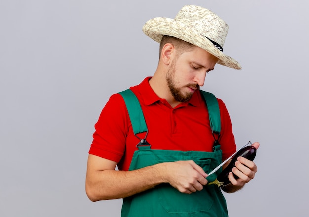 Junger hübscher slawischer Gärtner in der Uniform und im Hut, die Aubergine halten und es mit Bandmeter isoliert messen