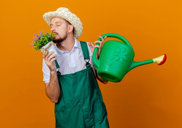 Junger hübscher slawischer Gärtner in Uniform und Hut, der Blumentopf und Bewässerung hält, kann Blumen mit geschlossenen Augen schnüffeln, die auf orange Wand lokalisiert werden