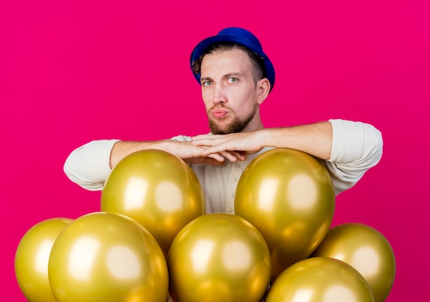 Junger hübscher slawischer Party-Typ, der Partyhut trägt, der hinter Luftballons steht, die Kamera betrachten, die Hände zusammenhalten, die Kussgeste tun, die auf purpurrotem Hintergrund lokalisiert wird