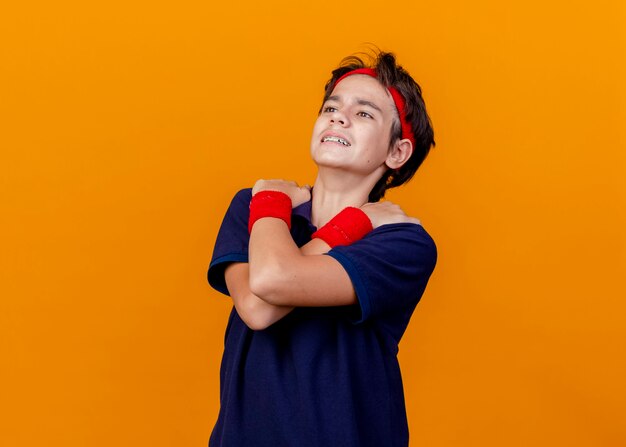 Junger hübscher sportlicher Junge, der Stirnband und Armbänder mit Zahnspangen trägt, die gerade halten, die Hände gekreuzt halten auf Schultern lokalisiert auf orange Hintergrund mit Kopienraum