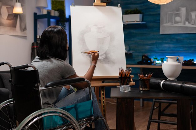Junger Illustrator im Rollstuhl zeichnet künstlerische Vasenillustration mit Grafikbleistift während des Malunterrichts im Kreativitätsstudio. Künstlerstudent mit Behinderung, der auf Leinwand zeichnet