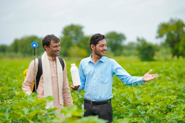 Junger indischer agronom, der dem landwirt eine flüssigdüngerflasche gibt und produktinformationen auf dem grünen landwirtschaftsfeld sagt.