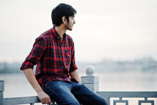 Junger indischer Student in kariertem Hemd und Jeans sitzt auf Handläufen am See