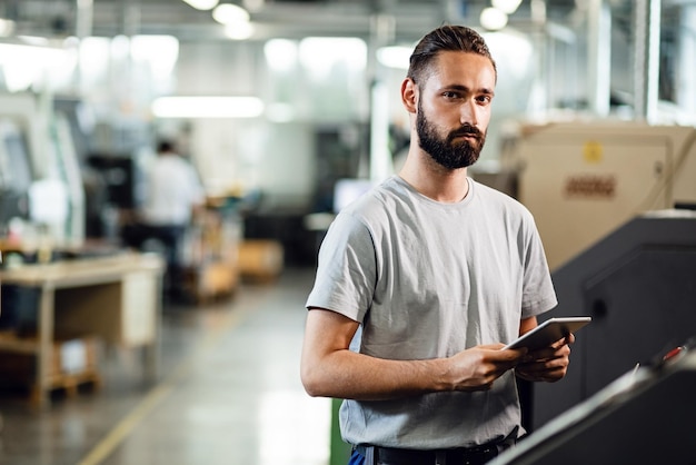 Junger Ingenieur, der ein Touchpad verwendet, während er eine CNC-Maschine in einer Fabrik bedient und in die Kamera schaut