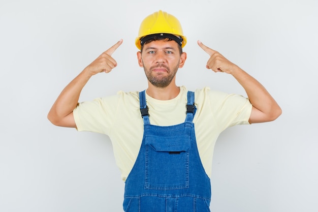 Junger ingenieur, der in uniform auf seinen helm zeigt und selbstbewusst aussieht, vorderansicht.