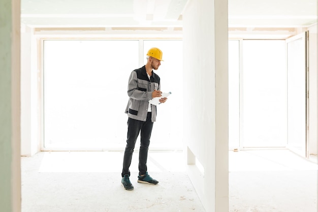 Junger Ingenieur in Arbeitskleidung und gelbem Helm, der den Plan neuer Wohnungen hält und nachdenklich auf die Wand schaut, mit großem Fenster im Hintergrund