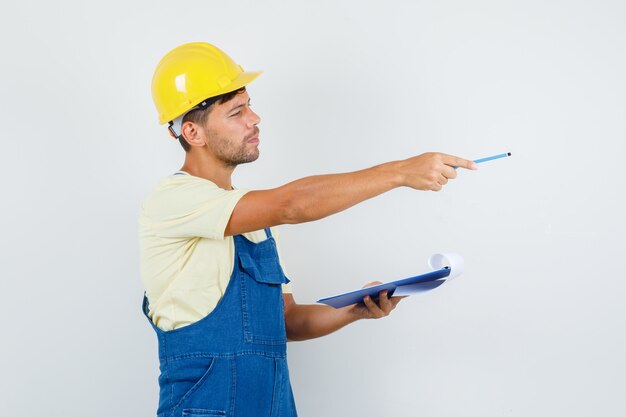 Junger Ingenieur in der Uniform, die Zwischenablage hält und zur Seite zeigt, Vorderansicht.