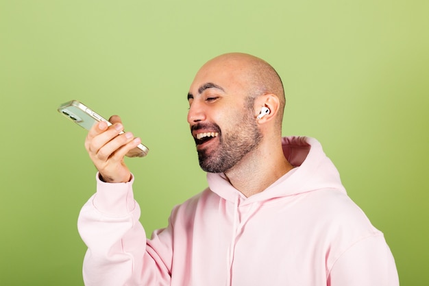Kostenloses Foto junger kahler kaukasischer mann in rosa kapuzenpulli isoliert, halten telefon glücklich positive aufnahme audio-nachricht
