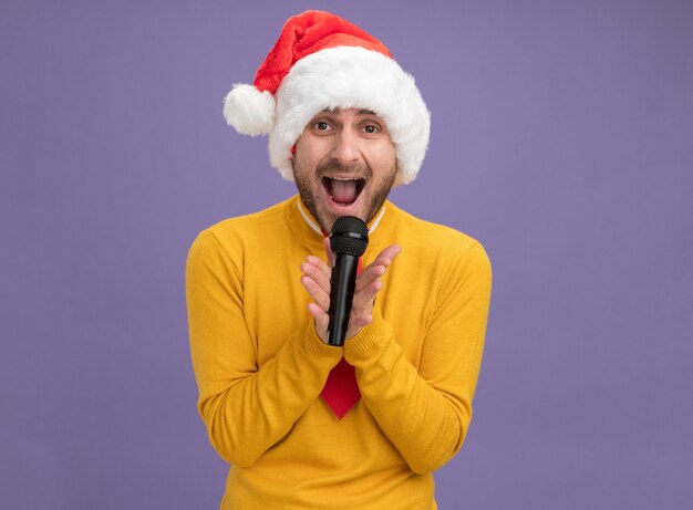 Junger kaukasischer Mann, der Weihnachtshut und Krawatte hält, die Mikrofon hält, das Kamera singt lokalisiert auf lila Hintergrund mit Kopienraum betrachtet