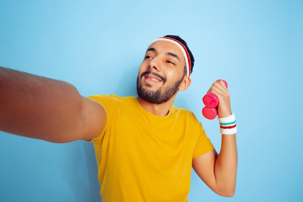 Junger kaukasischer Mann im hellen Kleidertraining auf blauem Raum Konzept des Sports, der menschlichen Gefühle, des Gesichtsausdrucks, des gesunden Lebensstils, der Jugend, des Verkaufs