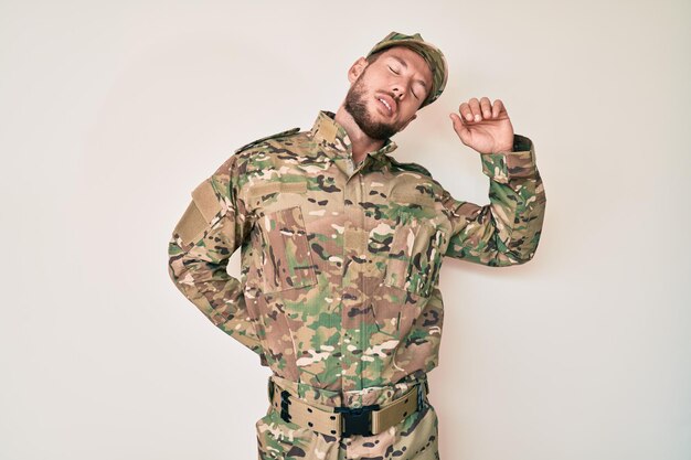 Junger kaukasischer Mann in Camouflage-Armeeuniform, der sich zurückstreckt, müde und entspannt, schläfrig und gähnend für den frühen Morgen