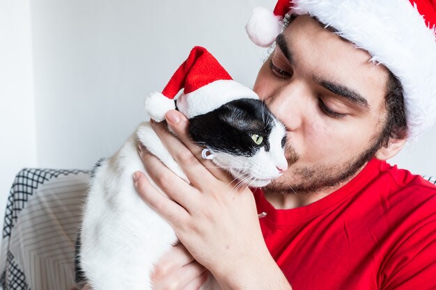 Junger kaukasischer Mann in einem Weihnachtsmannhut, der mit seinem weißen Schwarz und Katze küsst
