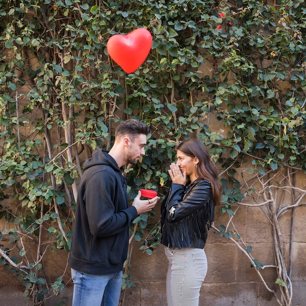 Kostenloses Foto junger kerl, der geschenkbox überraschter dame nahe fliegendem ballon in der form des herzens darstellt