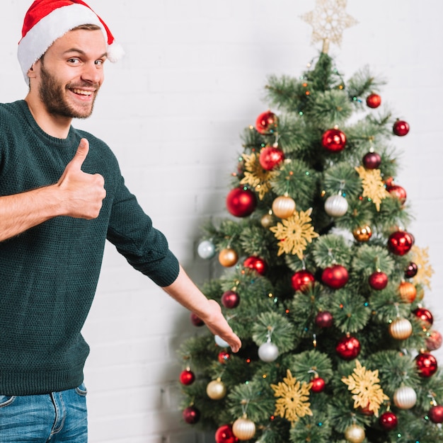 Junger Kerl, der guten nahen Weihnachtsbaum zeigt