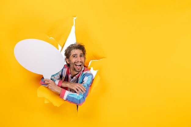 Junger Kerl, der weißen Ballon hält und in einem zerrissenen Loch und freiem Hintergrund in gelbem Papier lächelt
