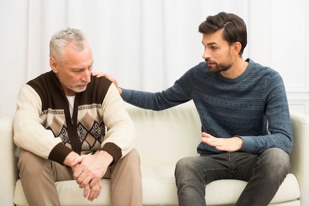 Junger Kerl mit der Hand auf Schulter des gealterten traurigen Mannes auf Sofa