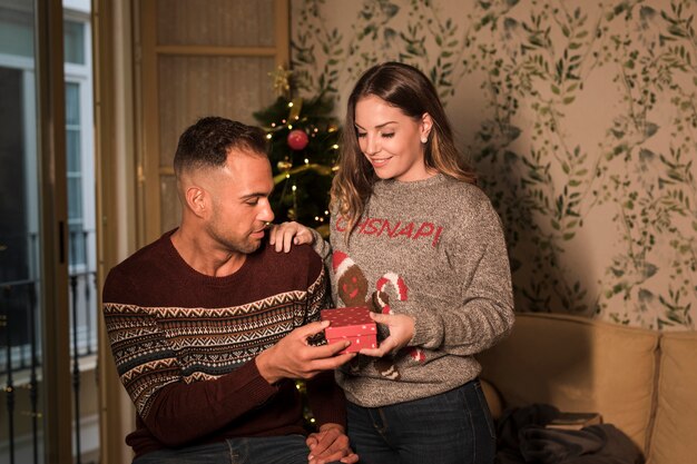 Junger Kerl mit Geschenkbox und fröhlicher Dame auf Sofa nahe Weihnachtsbaum