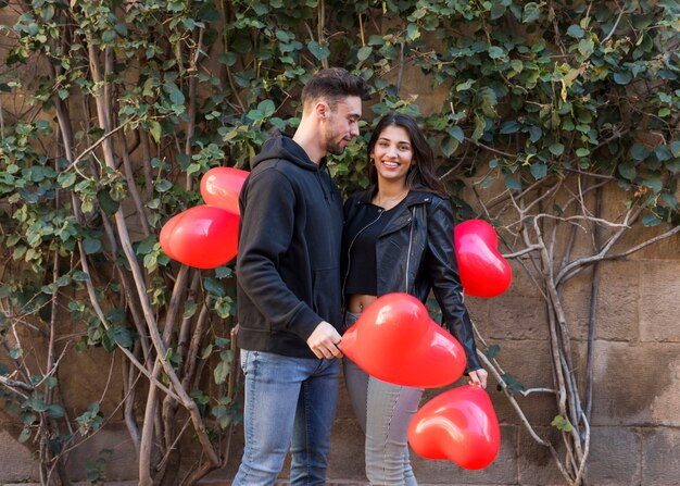 Junger Kerl nahe lächelnder Dame, die Ballone in der Form von Herzen hält