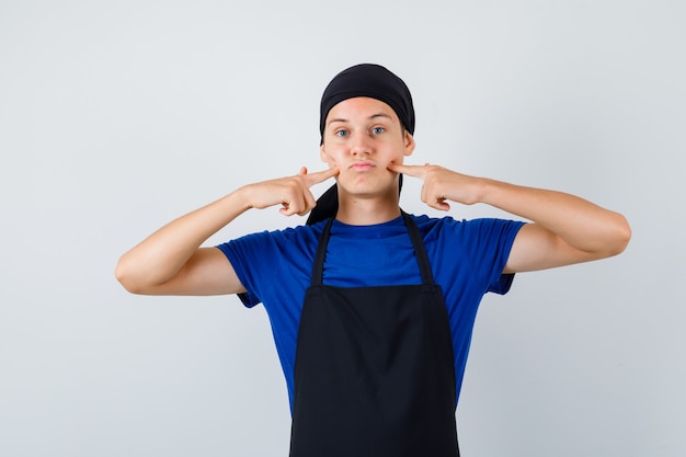 Junger Koch im T-Shirt, Schürze drückt Finger auf Grübchen und sieht gelangweilt aus, Vorderansicht.