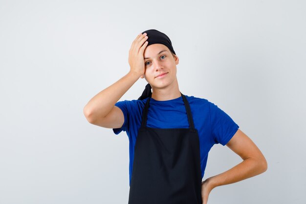 Junger Koch mit der Hand auf dem Kopf in T-Shirt, Schürze und fröhlichem Blick, Vorderansicht.