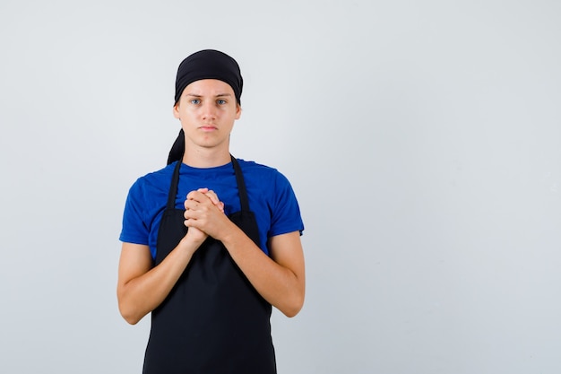 Junger Kochmann, der gefaltete Hände in flehender Geste in T-Shirt, Schürze zeigt und hoffnungsvoll aussieht, Vorderansicht.