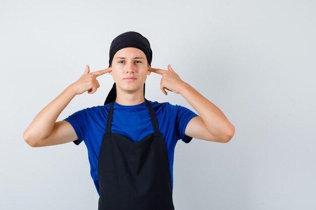 Junger Kochmann im T-Shirt, Schürze, die Ohren mit den Fingern verstopft und genervt aussieht, Vorderansicht.