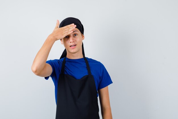 Junger Kochmann im T-Shirt, Schürze mit Hand auf der Stirn und vergesslicher Blick, Vorderansicht.